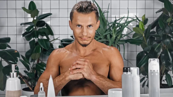 Hombre sin camisa con las manos apretadas mirando a la cámara cerca de plantas verdes sobre fondo borroso en el baño - foto de stock