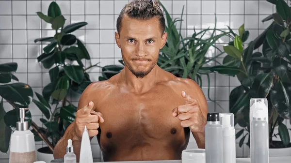 Homem sem camisa apontando com os dedos enquanto olha para a câmera perto de plantas verdes no fundo borrado no banheiro — Fotografia de Stock