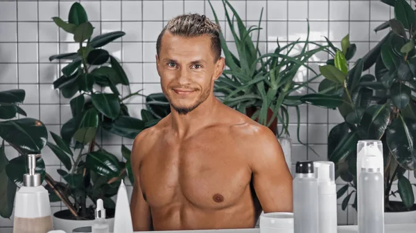 Hombre sonriente y sin camisa cerca de plantas verdes sobre fondo borroso en el baño - foto de stock