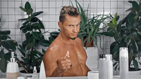 Young shirtless man pointing with finger while looking at camera near green plants on blurred background in bathroom — Stock Photo