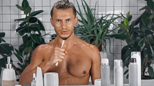 Мужчина без рубашки держит зубную щетку рядом с зелеными растениями на размытом фоне в ванной комнате — стоковое фото