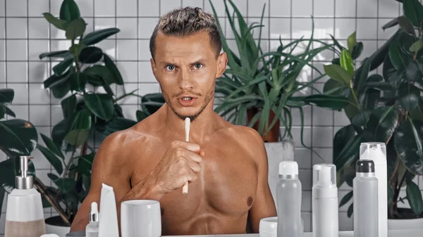 Homem sem camisa falando enquanto segurando escova de dentes perto de plantas verdes no fundo borrado no banheiro — Fotografia de Stock