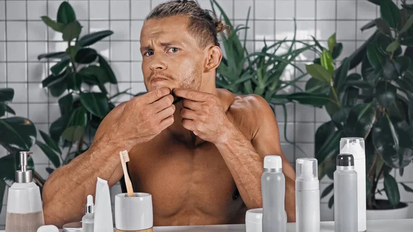 Homme torse nu pressant bouton sur le visage près de plantes vertes sur fond flou dans la salle de bain — Photo de stock