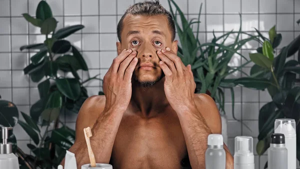 Junger Mann ohne Hemd berührt Gesicht in der Nähe grüner Pflanzen auf verschwommenem Hintergrund im Badezimmer — Stockfoto