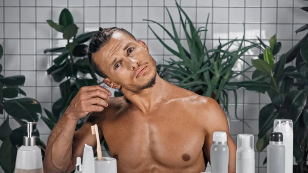 Мужчина без рубашки засовывает ватный тампон в ухо рядом с растениями на размытом фоне в ванной комнате — стоковое фото