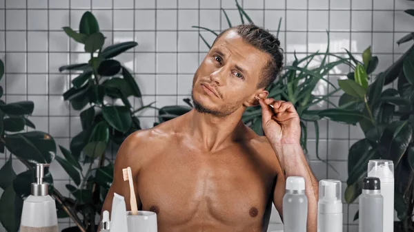 Молодой человек без рубашки засовывает ватный тампон в ухо рядом с растениями на размытом фоне в ванной комнате — стоковое фото
