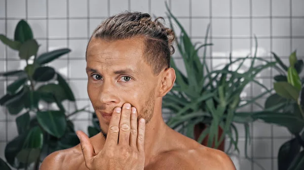 Bärtiger und hemdsloser Mann berührt Gesicht, während er im Badezimmer in die Kamera schaut — Stockfoto