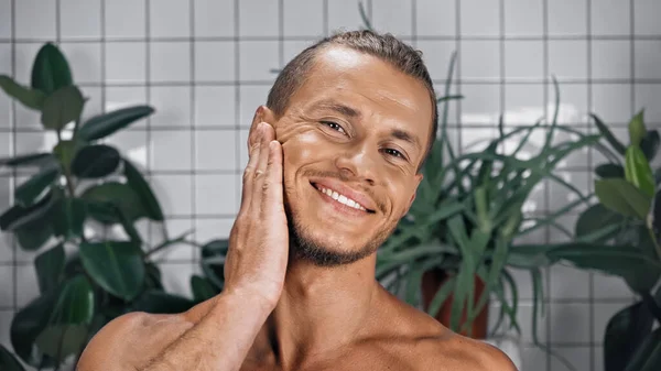 Позитивный мужчина трогает лицо и смотрит в камеру в ванной комнате — стоковое фото