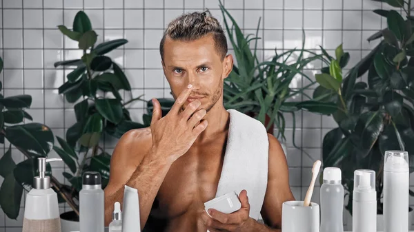 Homem barbudo aplicando creme facial no banheiro com plantas no fundo borrado — Fotografia de Stock