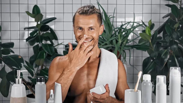 Homme heureux appliquant crème visage sur le nez dans la salle de bain avec des plantes sur fond flou — Photo de stock