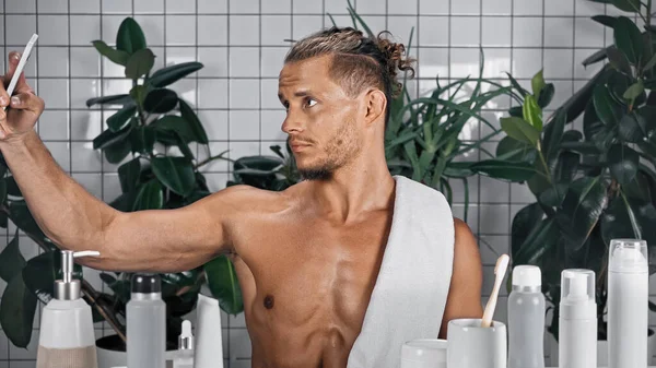 Hombre sin camisa tomando selfie en cuarto de baño con plantas sobre fondo borroso - foto de stock