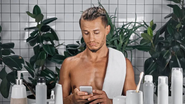 Homme torse nu utilisant smartphone dans la salle de bain avec des plantes sur fond flou — Photo de stock