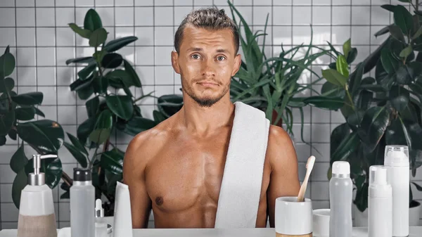 Uomo barbuto con asciugamano sul corpo senza maglietta in bagno vicino a piante su sfondo sfocato — Foto stock