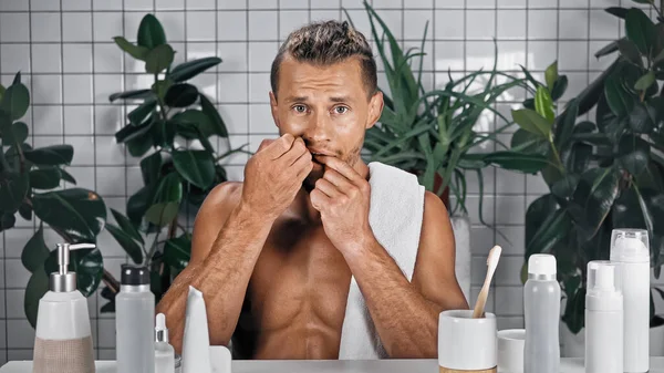 Hombre barbudo con toalla en el cuerpo sin camisa dientes de hilo dental en el baño cerca de las plantas sobre fondo borroso - foto de stock