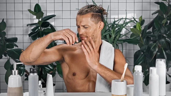 Uomo scontento con asciugamano sul corpo senza camicia gesticolando in bagno vicino a piante su sfondo sfocato — Foto stock