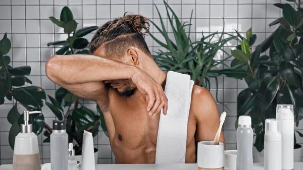 Мужчина с полотенцем на теле без рубашки нюхает подмышку в ванной рядом с растениями на размытом фоне — стоковое фото