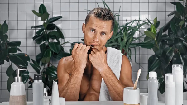 Bearded man with open mouth flossing teeth in bathroom near bottles and plants on blurred background — Stock Photo