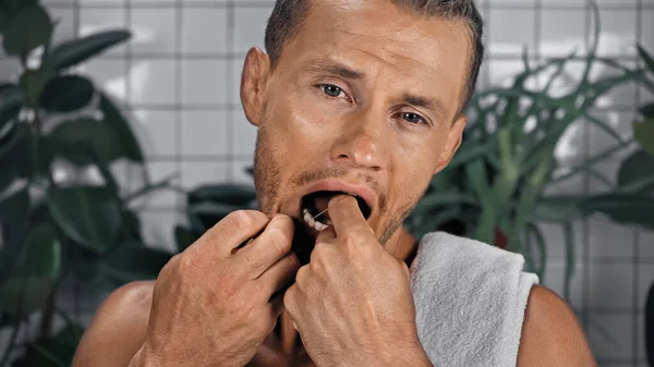 Homme avec bouche ouverte dents en soie dentaire dans la salle de bain près de plantes sur fond flou — Photo de stock
