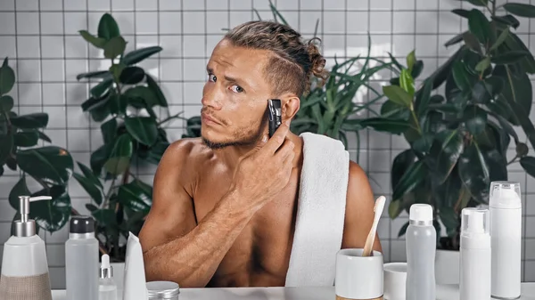 Hemdloser Mann mit Trimmer im Badezimmer in der Nähe von Flaschen und Pflanzen auf verschwommenem Hintergrund — Stockfoto