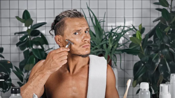 Hemdloser Mann mit Rasiermesser im Badezimmer in der Nähe von Flaschen und Pflanzen auf verschwommenem Hintergrund — Stockfoto