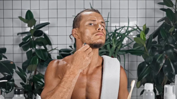 Homme barbu en utilisant un rasoir dans la salle de bain près des bouteilles et des plantes sur fond flou — Photo de stock