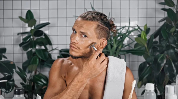 Shirtless man using razor in bathroom near bottles and green plants on blurred background — Stock Photo