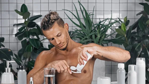 Безсоромний чоловік стискає зубну пасту на зубній щітці у ванній біля зелених рослин на розмитому фоні — стокове фото