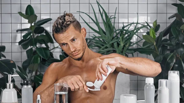 Hombre barbudo apretando pasta de dientes en el cepillo de dientes en el baño cerca de plantas verdes sobre fondo borroso - foto de stock