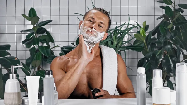Uomo senza camicia che applica schiuma da barba sul viso vicino a bottiglie e piante verdi su sfondo sfocato — Foto stock
