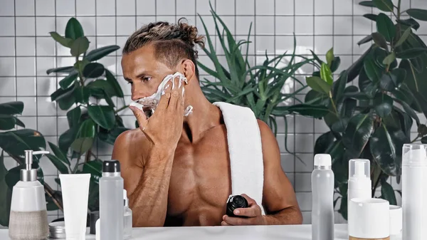 Homem sem camisa aplicando espuma de barbear no rosto perto de garrafas e plantas no fundo borrado — Fotografia de Stock
