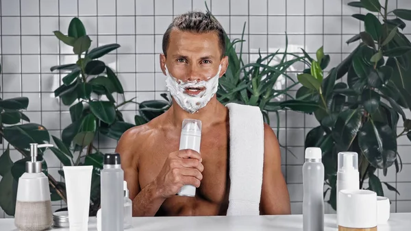 Мужчина без рубашки с пеной для бритья на лице держит бутылку рядом с растениями на размытом фоне — стоковое фото