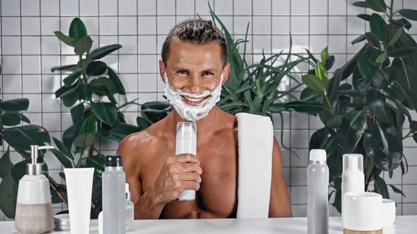 Homem feliz com espuma de barbear no rosto segurando garrafa perto de plantas no fundo borrado — Fotografia de Stock