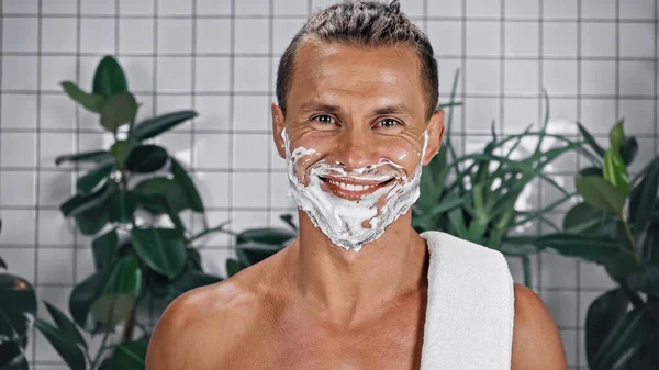 Joyful man with shaving foam on face looking at camera near plants on blurred background — Stock Photo