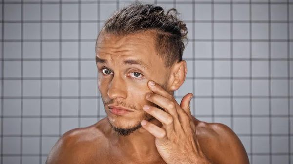 Бородатый мужчина трогает кожу возле глаза, смотрит в камеру в ванной комнате. — стоковое фото