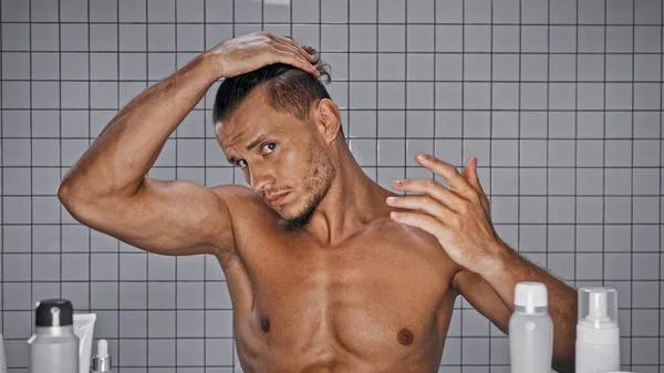 Без сорочки чоловік регулює волосся біля пляшок у ванній — стокове фото