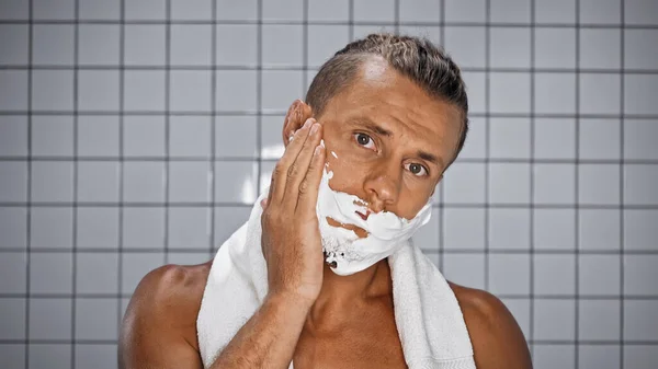 Bärtiger Mann blickt in Kamera und trägt Rasierschaum im Badezimmer auf — Stockfoto