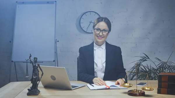 Lächelnder Versicherungsvertreter mit Blick auf Kamera in der Nähe von Geräten, Justizstatuette und Waage im Büro — Stockfoto