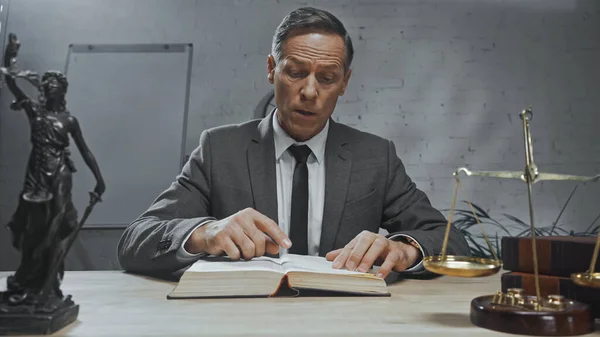 Un agent d'assurance lisant un livre près d'une statuette de justice et des balances au premier plan flou sur la table — Photo de stock
