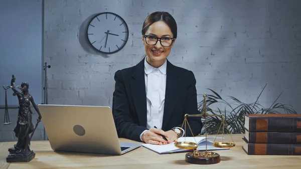 Agente de seguros sonriente sosteniendo pluma cerca de portátil, portátil y básculas en la mesa — Stock Photo