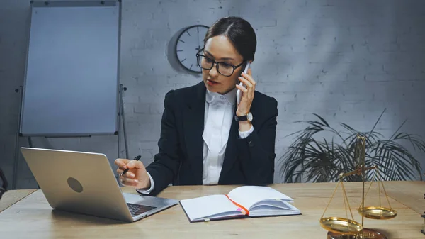 Agent d'assurance parlant sur smartphone près d'un ordinateur portable, ordinateur portable et balances sur la table — Photo de stock