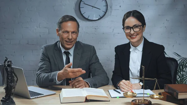 Agente de seguros sonriente apuntando con la mano cerca de colega, libros y computadora portátil en la mesa - foto de stock