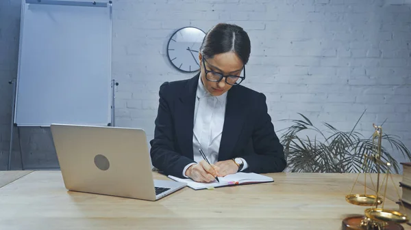 Agente de seguros escrevendo no notebook perto de laptop e escalas em primeiro plano desfocado — Fotografia de Stock