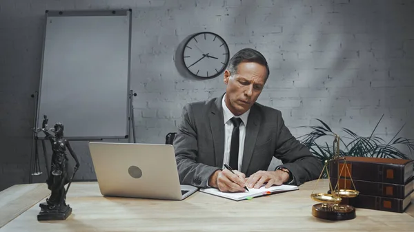 Versicherungsvertreter im Anzug schreibt auf Notizbuch neben Laptop, Bücher und Waagen auf Tisch — Stockfoto