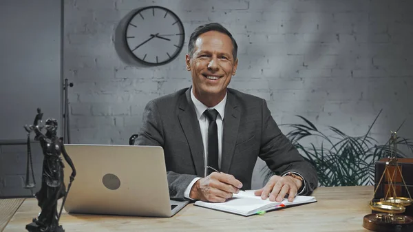 Smiling insurance agent looking at camera near notebook, laptop and statuette of justice on table — Stock Photo