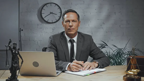 Versicherungsvertreter in offizieller Kleidung hält Stift in der Nähe von Notizbuch, Laptop und Justizstatuette auf verschwommenem Vordergrund — Stockfoto