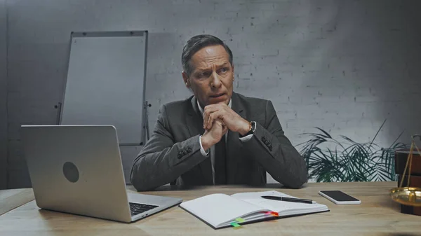 Agente de seguros reflexivo mirando hacia otro lado cerca de dispositivos, cuaderno y básculas en la mesa de trabajo - foto de stock