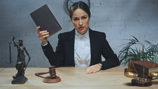 Versicherungsvertreter hält Buch in der Hand, während er in die Kamera in der Nähe von Hammer, Statuette der Gerechtigkeit und Waage auf dem Tisch blickt — Stockfoto