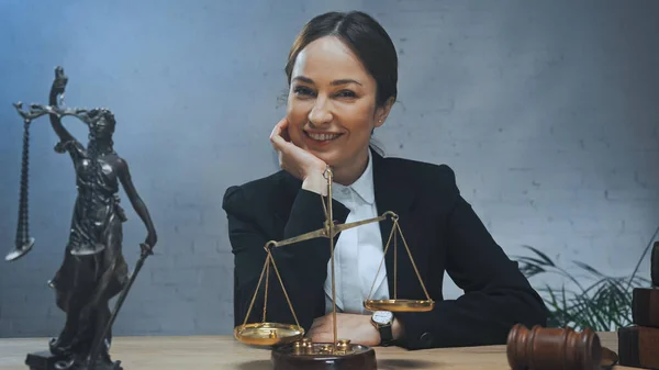 Smiling insurance agent looking at camera near statuette of justice, scales and gavel on blurred foreground — Stock Photo