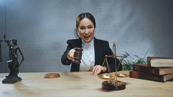 Lächelnder Versicherungsvertreter mit Hammer in der Nähe von Büchern, Waagen und Justizstatuette auf dem Tisch — Stockfoto