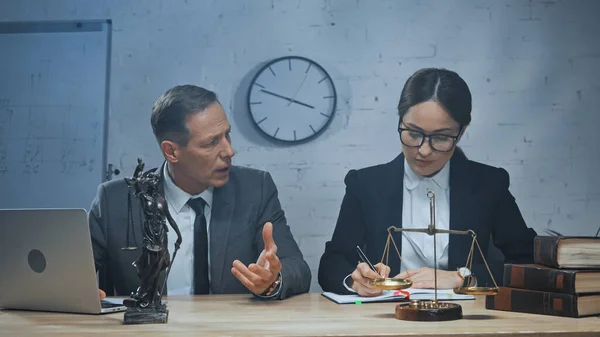 Agente de seguros hablando con un colega escribiendo en un cuaderno cerca de la computadora portátil, libros y balanzas en la mesa - foto de stock